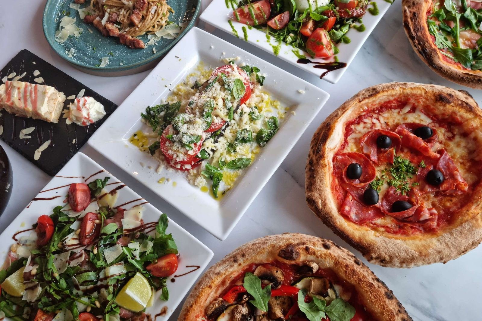 Een marmeren tafel met twee pizza&rsquo;s uit de houtoven, pasta, salades met tomaat en rucola en een dessert.